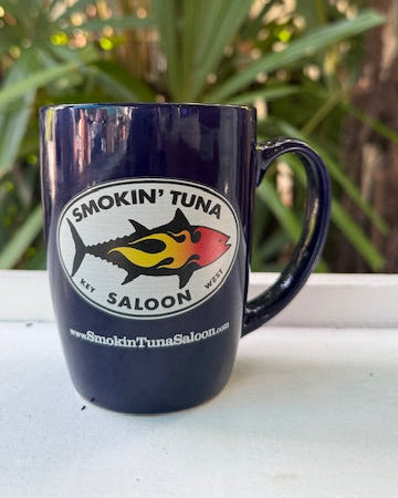 Navy blue mug with 'Smokin' Tuna Saloon' logo on a white surface with green foliage in the background